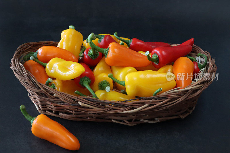 一堆红色、橙色和黄色的迷你甜椒(Capsicum annuum)的特写图片，绿色的茎放在柳条篮子里，黑色背景，重点放在前景上，复制空间